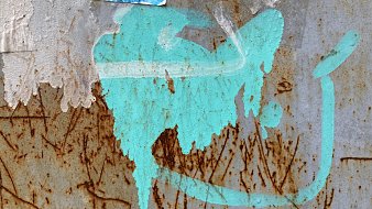 L'image présente une surface en métal, probablement rouillée, avec des marques et des textures variées. On y voit une tache de couleur turquoise qui semble avoir été projetée. Cette tache a une forme fluide et irrégulière, évoquant une sorte de mouvement ou de dynamisme. À proximité, on peut distinguer des traces brunes, résultant de la rouille, qui ajoutent un contraste marqué à la couleur vive. Le fond est texturé, avec des stries et des imperfections, suggérant l'âge et l'usure du matériau. L'ensemble crée une atmosphère à la fois urbaine et artistique.