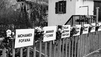L'image montre une scène en noir et blanc, où l'on aperçoit une maison avec un balcon en bois. Devant la maison, il y a une clôture sur laquelle sont fixés plusieurs panneaux blancs. Chaque panneau présente un prénom et un nom, probablement en hommage à des personnes. Des fleurs blanches sont disposées autour de chaque panneau, ajoutant une touche de douceur. En arrière-plan, on peut deviner des arbres et la nature environnante, suggérant un cadre paisible et naturel.