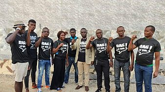 L'image montre un groupe de onze personnes rassemblées, certaines portant des t-shirts noirs avec des inscriptions. Ils affichent tous des expressions déterminées et se tiennent debout, avec quelques-uns levant le poing en signe de solidarité ou de protestation. Les membres du groupe sont de différents sexes et portent des vêtements variés, allant de t-shirts décontractés à des vêtements plus formels. L'arrière-plan est constitué d'un mur en pierre, ajoutant une texture neutre à la scène. L'ambiance semble énergique et engagée.