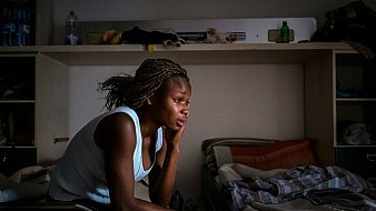 L'image dépeint une femme assise sur le bord d'un lit dans une pièce sobre. Elle a une coiffure tressée et porte un haut sans manches de couleur claire. Son expression est pensive, montrant une profonde réflexion ou peut-être de la tristesse. La lumière douce qui filtre à travers une fenêtre éclaire son visage, créant des ombres sur ses traits. La chambre semble être en désordre, avec des objets éparpillés et des draps non rangés. L'atmosphère de l'image évoque un sentiment de solitude et de contemplation.