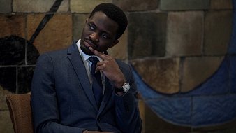 L'image montre un homme assis dans un environnement intérieur. Il porte un costume sombre avec une chemise claire et une cravate, ce qui dénote un certain formalisme. Son expression est pensive, avec un doigt sur le menton, suggérant qu'il réfléchit à quelque chose de sérieux. En arrière-plan, on aperçoit un mur aux motifs colorés, avec des formes géométriques qui ajoutent une touche artistique à la scène. L'ambiance est calme et réfléchie.