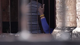 Cette image montre une scène à l'intérieur d'un espace confiné, avec des barreaux visibles au premier plan. On distingue des silhouettes humaines derrière les barreaux. Une main, portant un bracelet, s'élève légèrement, témoignant d'un geste de communication ou de contact. Les murs autour des barreaux semblent anciens et en pierre, ajoutant un élément d'authenticité à l'environnement. L'éclairage suggère que la scène se déroule à l'extérieur, avec une lumière naturelle qui éclaire partiellement l'espace.