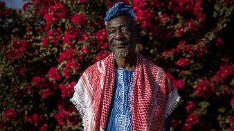 L'image montre un homme debout devant un fond vibrant de fleurs roses, probablement des bougainvilliers. Il porte une tenue traditionnelle riche en couleurs, avec un tissu bleu et des motifs complexes. Sur ses épaules, il a une écharpe ou un châle rouge et blanc, typique de certaines cultures. Son visage exprime sagesse et dignité, avec des traits marqués par le temps. Le contraste entre ses vêtements et les fleurs éclatantes crée une atmosphère chaleureuse et accueillante.