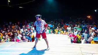 L'image montre une scène vivante et animée. Au premier plan, un artiste, habillé de manière colorée avec une chemise à motifs et un short, se tient sur une scène en bois. Son expression enthousiaste et son attitude dynamique suggèrent qu'il est en plein spectacle. Derrière lui, une foule nombreuse est présente, composée de personnes de tous âges et de toutes origines, certaines souriant et d'autres captivées par la performance. Les lumières autour de la scène créent une atmosphère festive, tandis que l'obscurité en arrière-plan met l'accent sur l'énergie du moment. Les spectateurs semblent engager et réceptifs, témoignant de l'ambiance joyeuse d'un événement en plein air.