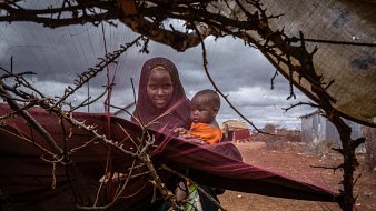 L'image montre une femme et un enfant au sein d'un environnement difficile. La femme, vêtue d'un voile sombre et d'habits traditionnels, tient l'enfant dans ses bras. L'enfant, portant un vêtement orange, semble curieux et regarde autour de lui. Ils sont entourés de branches et de tissus, ce qui forme une sorte d'abri rudimentaire. Le sol est poussiéreux et l'atmosphère est chargée d'une lumière diffuse, suggérant un ciel nuageux. Cette scène évoque la résilience et le lien fort entre la mère et son enfant dans un contexte de précarité.