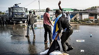 L'image montre une scène de manifestation dans une rue. Au premier plan, on peut voir des personnes en mouvement, certaines en train de gesticuler, tandis qu'une personne est à terre, apparemment blessée. L'un des manifestants porte un drapeau, probablement symbolique. En arrière-plan, il y a un véhicule de police, ce qui suggère une présence des forces de l'ordre. Le sol est couvert de débris, comme des affiches et des bouteilles, témoignant de la tension de la situation. L'ambiance semble intense et sérieuse, avec une impression de conflit.