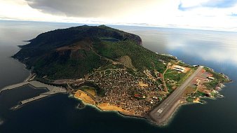 Imagine une île isolée au milieu d'une vaste étendue d'eau calme et scintillante. La surface de l'eau reflète un ciel nuageux, ce qui donne une ambiance sereine. Sur l'île, il y a une montagne verdoyante qui se dresse majestueusement, recouverte de végétation dense et luxuriante. Au bas de la montée, on aperçoit un village coloré, composé de petites maisons accolées, créant un patchwork de teintes. Les bâtiments semblent être en harmonie avec la nature environnante. Sur la côte, il y a un port avec des structures en pierre, probablement destinées à accueillir des bateaux. À l'extrémité de l'île, une piste d'atterrissage est visible, signalant une certaine accessibilité même dans ce coin reculé du monde. L'ensemble évoque un mélange de tranquillité, de vie communautaire et de beauté naturelle.