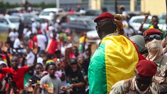L'image montre une scène dynamique et pleine d'énergie. Au premier plan, un homme en uniforme militaire, portant un béret rouge, se tient sur une plateforme surélevée. Il est enveloppé dans un drapeau aux couleurs vives du Mali, qui est vert, jaune et rouge. Autour de lui, une foule enthousiaste s'agite, brandissant des drapeaux et exprimant leur joie. Les visages des personnes dans la foule montrent des émotions variées allant de l'excitation à la fierté. En arrière-plan, on peut voir des voitures stationnées et d'autres participants se mêlant dans une atmosphère de célébration collective. L'ensemble dégage un fort sentiment d'unité et de patriotisme.