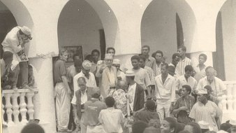 L'image représente une scène de rassemblement en extérieur, probablement lors d'un événement communautaire ou historique. Au premier plan, un groupe de personnes se regroupe autour d'une figure centrale, tandis qu'une autre personne, en haut à gauche, semble prendre une photo. Les participants portent des vêtements variés, certains avec des chapeaux. L'architecture en arrière-plan témoigne d'un style colonial, avec des arches et des balustrades. L'ambiance générale paraît vivante et joyeuse, suggérant un moment d'unité et de partage au sein d'une communauté.