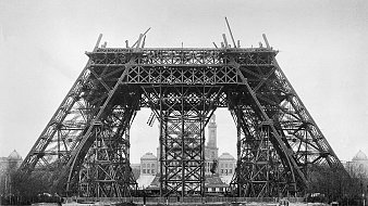 Cette image montre une phase de construction de la célèbre Tour Eiffel à Paris. On peut voir une structure métallique imposante, caractérisée par des poutres en fer entrecroisées. La tour, encore incomplète, présente une base large et stable avec des arcades qui s'élèvent vers le ciel. À l'arrière-plan, on distingue un bâtiment, peut-être une mairie, qui ajoute un contexte urbain à la scène. L'ambiance globale de l'image est monochrome, ce qui renforce l'aspect historique et donne un aperçu des débuts de cette icône de l'architecture. Le sol est encore en chantier, avec des débris et des outils, ce qui rappelle le processus de construction en cours.