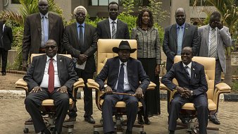 L'image présente un groupe de personnes posant ensemble à l'extérieur d'un bâtiment. Au centre, on voit trois hommes assis dans de grandes chaises confortables. Le premier homme à gauche porte des lunettes, un costume sombre et une cravate rouge. Le second, au centre, est vêtu d'un costume noir, d'une cravate et d'un chapeau noir, et semble être le leader du groupe. Le troisième homme, à droite, porte également un costume et une cravate. Derrière eux, plusieurs personnes se tiennent debout, toutes en tenue formelle. L'environnement est verdoyant, avec des plantes et un chemin en pavé. L'atmosphère semble sérieuse et officielle, suggérant une rencontre importante.