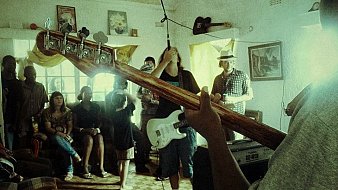 L'image montre une scène musicale vivante dans une pièce chaleureuse. À l'avant, on voit un musicien tenant une basse, tandis qu'un autre, avec une guitare électrique, se concentre sur sa performance. Les membres du groupe sont entourés d'un public qui semble apprécier la musique. Les visages sont tournés vers les musiciens, certains se balançant au rythme. La pièce est décorée avec des tableaux aux murs et des rideaux qui laissent filtrer une lumière douce. L'ambiance est conviviale, remplie d'énergie et de passion pour la musique.