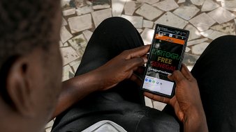 Sur cette image, on voit une personne assise, tenant un smartphone dans ses mains. La personne semble attentive à l'écran, qui affiche un site ou une application avec les mots "CARTOGR" en gros caractères, suivi de "FREE SENEGAL". Le fond est constitué de carreaux de mosaïque, tandis que la personne porte un t-shirt clair et un pantalon noir. L'arrière-plan et la position des mains indiquent une ambiance calme et concentrée.