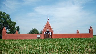 L'image présente une grande entrée de style architectural distinctif, peinte en rouge, qui s'élève au-dessus d'un champ de maïs. La structure est dotée d'un mur haut, orné de pointes qui se projettent vers le ciel, créant un aspect à la fois imposant et décoratif. Au centre, une porte en fer forgé permet d'entrer, et on peut apercevoir un drapeau français flottant au-dessus. Le ciel d'un bleu éclatant, parsemé de nuages légers, contraste avec la verdure des jeunes plants de maïs qui s'étendent devant le mur, ajoutant une touche de fraîcheur à la scène.