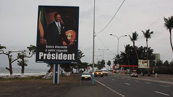 L'image montre un grand panneau publicitaire sur le bord d'une route. Ce panneau présente le portrait d'un homme en costume, avec un air solennel, tenant un insigne ou un symbole de pouvoir. En dessous de son image, il y a un texte qui dit : "Gloire éternelle à notre regretté Président", exprimant un hommage à un dirigeant décédé. En arrière-plan, on peut apercevoir une route avec quelques voitures et des palmiers, ainsi qu'une plage visible à proximité. Le ciel est nuageux, ce qui donne une ambiance mélancolique à la scène.