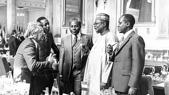 L'image représente un groupe de cinq hommes en discussion dans un cadre formel, probablement lors d'une conférence ou d'une réunion. Ils sont habillés en costumes élégants, dont certains sont accompagnés de cravates. Les visages montrent une interaction animée, avec des gestes de mains qui suggèrent une conversation engageante. En arrière-plan, on aperçoit des drapeaux, ce qui indique un contexte international. La scène se déroule dans une grande salle, ornée et bien éclairée, ajoutant une atmosphère de sérieux et de respect.