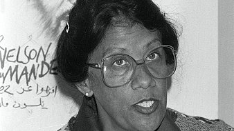 L'image montre une femme avec des lunettes épaisses, au visage expressif et aux cheveux noirs coiffés en un chignon. Elle porte un pull coloré avec un motif fleuri sur fond clair. En arrière-plan, on aperçoit une illustration d'un homme, probablement un portrait, qui ajoute un contexte à la scène. L'atmosphère semble sérieuse, et la femme semble engagée dans une discussion ou un discours. Ses traits et son regard transmettent une certaine détermination et une passion pour le sujet abordé.