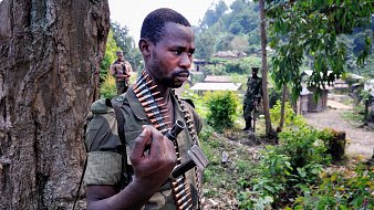 L'image montre un soldat debout près d'un arbre, portant un uniforme militaire camouflage. Il a une ceinture de munitions autour de son torse, et il tient une arme à la main. Son expression est sérieuse, indiquant la concentration et la vigilance. En arrière-plan, d'autres soldats sont visibles, et le paysage est verdoyant, avec des habitations simples au loin. L'atmosphère dégage une impression de tension en raison du contexte militaire.