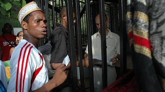 L'image montre un groupe de personnes rassemblées autour d'une structure métallique, probablement une grille ou une clôture. Un jeune homme au premier plan, portant un maillot blanc avec des rayures rouges et un chapeau traditionnel, exprime une certaine agitation ou curiosité. À côté de lui, d'autres personnes, dont un homme en chemise claire, semblent discuter ou échanger des documents. L'environnement est extérieur et verdoyant, suggérant un lieu fréquenté. Les expressions des visages indiquent une interaction dynamique et engageante entre les personnes présentes.