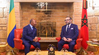 L'image montre deux hommes assis face à face dans un salon chaleureux et élégant. À gauche, un homme en costume bleu, avec une cravate, est légèrement incliné vers l'avant, écoutant attentivement. À droite, un autre homme porte un costume à carreaux bleu foncé et une cravate assortie, affichant une expression sérieuse tout en participant à la conversation. Derrière eux, on peut voir un mur en briques claires et une cheminée décorative. Il y a également des éléments de décoration comme des fauteuils rouges et un tableau avec un drapeau marocain sur le côté droit. Une lumière douce illumine la pièce, créant une atmosphère conviviale.