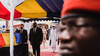 L'image montre une scène d'événement officiel, probablement une cérémonie. Au premier plan, il y a un homme portant des lunettes et un uniforme sombre, dont le visage est flou mais qui semble attentif. En arrière-plan, deux autres hommes, l'un en costume noir et l'autre en tenue traditionnelle blanche, marchent sous un chapiteau aux couleurs vives de jaune, rouge et bleu. Le chapiteau est décoré de tissu et crée une atmosphère formelle. La foule semble nombreuse et attentive, rendant l'ensemble de la scène solennelle et respectueuse. L'éclairage semble naturel, suggérant peut-être une belle journée ensoleillée.
