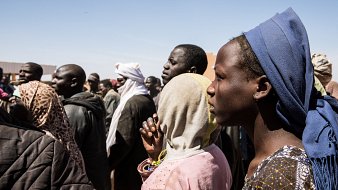 L'image montre un groupe de personnes se tenant debout, la plupart tournées vers un même point. On peut distinguer des hommes et des femmes de différents âges, habillés de vêtements variés, souvent colorés et traditionnels. Certaines personnes portent des couvertures sur la tête ou des scarves. L'expression sur leurs visages semble sérieuse, indiquant peut-être une attente ou une attention particulière. L'arrière-plan présente un ciel clair, suggérant une journée ensoleillée, et des structures émergent faiblement en arrière-plan, ajoutant à l'environnement d'un rassemblement. Le moment semble chargé d'émotions et de tension collective.