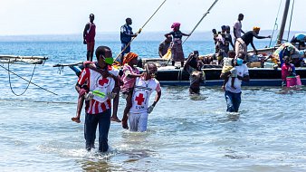 L'image montre une scène animée au bord de la mer. On peut voir plusieurs personnes dans l'eau peu profonds, certaines portant des vêtements colorés. Des hommes et des femmes s'affairent autour de petites embarcations. Certaines personnes transportent des enfants, tandis que d'autres semblent s'entraider. En arrière-plan, il y a une barque avec des passagers qui attendent. L'ambiance semble être pleine de vie et de solidarité, avec un ciel dégagé et une mer calme.