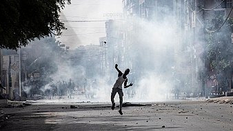 L'image dépeint une scène urbaine troublée, où la tension est palpable. Au premier plan, une silhouette humaine, probablement un homme, est en mouvement, lançant un projectile. Il semble néanmoins être entouré de foules, suggérant une situation tumultueuse. Des nuages de fumée épaisse s'élèvent dans l'air, créant une atmosphère de chaos, tandis que des débris jonchent le sol, témoignant d'une agitation récente. En arrière-plan, on peut deviner des bâtiments urbains flous, et la lumière est sombre, renforçant l'impression d'une émeute ou d'une protestation. Les sons et odeurs d'une ville agitée pourraient également accompagner cette image, avec des cris, des sirènes, ou l'odeur de fumée.