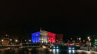 Imaginez un bâtiment majestueux illuminé par des lumières tricolores. La façade est peinte de bleu, de blanc et de rouge, évoquant les couleurs du drapeau français. Le ciel nocturne est sombre, créant un contraste saisissant avec les lumières vibrantes. À l'avant, vous pouvez sentir la présence d'une place animée, avec des voitures qui circulent et des lampadaires qui diffusent une lumière douce sur le pavé. La scène dégage une ambiance festive et patriotique, symbolisant la fierté nationale.