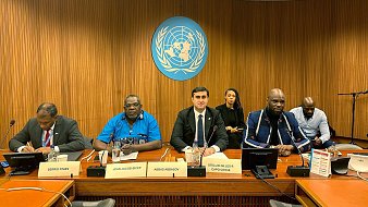 L'image montre une scène à une table de conférence, probablement lors d'une réunion aux Nations Unies. Au centre, un homme en costume noir et cravate blanche, avec un air sérieux, semble être le porte-parole ou le modérateur. À sa gauche, un homme portant une chemise bleue et des lunettes, avec un air attentif. À sa droite, un homme d'apparence plus décontractée avec un pull et une barbe, qui semble également participer à la discussion. En arrière-plan, plusieurs personnes sont assises, écoutant attentivement. Le décor est sobre, avec des murs en bois et le logo des Nations Unies en arrière-plan.