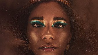 L'image montre un portrait d'une femme avec une peau sombre et un regard intense. Elle porte une couronne dorée ornée de serpents. Son maquillage se distingue par des teintes vibrantes de turquoise autour des yeux, ajoutant un contraste frappant à son visage. La lumière douce crée une ambiance mystérieuse, tandis qu'un brouillard léger autour d'elle donne une impression d'éthéré. L'ensemble dégage une aura de majesté et de puissance.