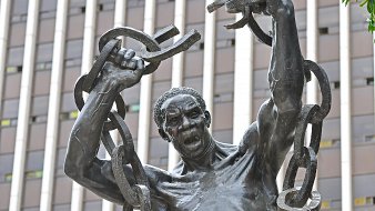Cette image représente une statue en bronze d'un homme robuste. Il se tient debout, les bras levés au-dessus de sa tête, brandissant de lourdes chaînes brisées, symbolisant la libération et la force. Son visage exprime une intense émotion, avec une bouche ouverte comme s'il poussait un cri de victoire ou de défi. La sculpture est placée devant un bâtiment moderne, avec des lignes verticales qui soulignent contraste entre le passé et le présent. L'œuvre évoque des thèmes de résilience et de lutte pour la liberté.