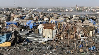 L'image montre un paysage de bidonville, où l'on peut distinguer des habitations précaires construites à partir de matériaux recyclés tels que des tôles, des bâches en plastique et des branches de bois. Ces structures sont souvent assez rudimentaires et forment des petites communautés. Le sol est jonché de déchets et de débris divers, reflétant une forte pollution. À l'arrière-plan, on aperçoit des bâtiments en dur, ainsi qu'une mosquée avec son minaret, indiquant la présence d'une ville proche. L'atmosphère dégage une impression de désolation, mais aussi de vie, avec des traces humaines visibles malgré les conditions difficiles.