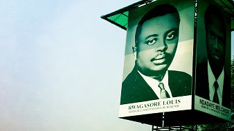 Cette image montre un grand panneau publicitaire ou une affiche située dans un environnement urbain. Sur le panneau, il y a un portrait en noir et blanc d'un homme, sous lequel est inscrit son nom : "RWAGASORE LOUIS", avec la mention "Héros de l'indépendance du Burundi". À côté de ce portrait, on aperçoit un autre visage (sans nom visible). Le fond de l'image présente un ciel légèrement brumeux et des bâtiments en arrière-plan, ce qui indique un paysage urbain. L'ambiance de l'image est à la fois historique et commémorative, mettant en avant des figures importantes pour le Burundi.