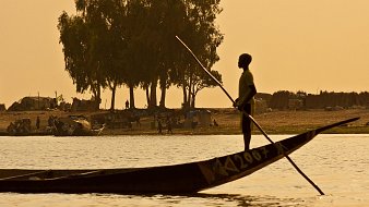 Imaginez une scène paisible au bord d'un fleuve. Un jeune garçon se tient debout dans une pirogue traditionnelle, les pieds ancrés sur le fond de la barque. Il utilise une longue perche pour diriger son embarcation à travers l'eau calme. En arrière-plan, on peut apercevoir un ruisseau de silhouettes d'arbres élancés, probablement des manguiers ou des sycomores, se découpant contre le ciel orangé du crépuscule. Sur la rive, des groupes de personnes s'affairent, tandis que des abris faits de toile et de paille sont visibles, suggérant une vie communautaire active. L'atmosphère est tranquille, teintée d'une chaleur douce, et l'ensemble dégage une sensation de sérénité et de connexion à la nature.