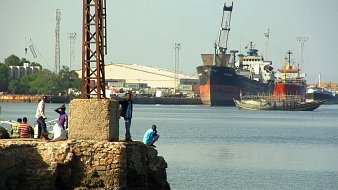 Imaginez un port animé. Au premier plan, il y a un quai en pierre avec plusieurs personnes : certaines sont debout, discutant, tandis que d'autres se penchent sur le bord pour observer l'eau. L'atmosphère est vivante, remplie d'échos de rires et de conversations. À l'arrière-plan, de grands navires sont amarrés, leurs coques visibles au-dessus de l'eau paisible. Des grues et des poteaux élancés se dressent, témoignant des activités portuaires. L'air sent légèrement le sel et le poisson, créant une ambiance maritime authentique.
