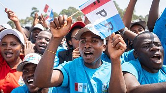 L'image montre un groupe de personnes en train de célébrer ou de soutenir une cause. Les participants portent des t-shirts bleus, et certains brandissent des drapeaux aux couleurs vives, portant l'inscription "FPR". Leur visage exprime de la passion et de l'enthousiasme, avec des sourires et des cris de joie. En arrière-plan, on aperçoit d'autres supporters, créant une atmosphère festive et animée. L'environnement semble ensoleillé, et l'énergie collective est palpable, illustrant un moment de solidarité et de camaraderie.