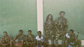 L'image montre un groupe de jeunes hommes, assis et debout, le long d'un mur vert. Les hommes portent des uniformes militaires camouflés pour certains, et d'autres sont habillés de manière plus décontractée. Ils semblent être dans un environnement extérieur, avec un sol en pierre. Quelques-unes des personnes tiennent des objets qui ressemblent à des sacs ou des équipements militaires. L'ambiance est tranquille, mais dégage une impression de camaraderie parmi le groupe.