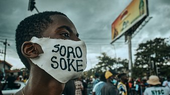 L'image montre une scène de manifestation dans une rue animée. Au premier plan, un jeune homme porte un masque blanc sur lequel est inscrit "SORO SOKE" en lettres noires, un slogan qui évoque la lutte pour les droits et la justice. Son regard est déterminé, et on peut sentir l'énergie de la foule qui l'entoure. À l'arrière-plan, des silhouettes de personnes se déplacent, et des affiches publicitaires peuvent être aperçues. Le ciel est nuageux, ce qui ajoute une atmosphère de tension à la scène. L'ensemble dégage une forte impression d'engagement et de solidarité.