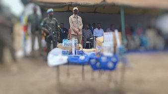 L'image montre un événement en plein air, avec une tente sur le côté. Au centre, on voit une table couverte d'objets et de brochures, probablement liés à une présentation ou un programme. À l'arrière-plan, plusieurs personnes sont assises, y compris des enfants et des adultes. Un homme se tient debout, portant une casquette et un masque, semblant être un intervenant ou une personne d'autorité. La scène reflète un moment de rassemblement communautaire, chargé d'une atmosphère d'échange et d'information.