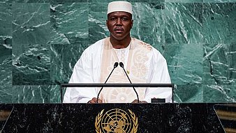 L'image montre un homme debout derrière un pupitre lors d'un discours. Il porte un costume traditionnel, probablement en blanc, avec un embellissement distinctif. Son teint est sombre et il a une expression sérieuse, indiquant qu'il est en train de s'adresser à un public. Le fond de l'image est constitué de panneaux de marbre vert, ce qui crée un contraste avec sa tenue. Le pupitre est orné du symbole des Nations Unies, représentant la mission internationale de paix et de coopération.