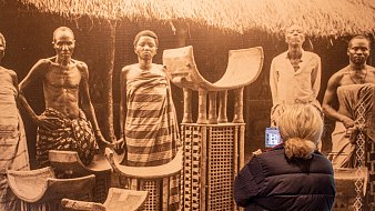 Dans cette image, on aperçoit une scène historique en noir et blanc montrant plusieurs personnes debout, semblant faire partie d'une communauté traditionnelle. Il y a des hommes et des femmes, vêtus de tissus authentiques, avec des poses qui expriment la dignité et la culture. Au centre, on voit des objets artisanaux, comme des chaises ou des tabourets, fabriqués en matériaux naturels, qui portent des motifs décoratifs. À l'arrière-plan, on devine une architecture simple, avec un toit de chaume, créant un cadre authentique. Une femme, de dos, observe cette scène, tenant un appareil dans ses mains, captivée par ce témoignage du passé. L'ambiance générale évoque un sentiment de respect pour les traditions et les histoires des personnes représentées.