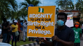 Sur l'image, une manifestation a lieu dans la rue. Un homme se tient au premier plan, portant un masque. Il tient une grande pancarte colorée qui affiche le message suivant : "#DIGNITY TO LIFE IS OUR Birth-Right Not A Privilege" en lettres grandes et noires sur un fond orange. En arrière-plan, on aperçoit un groupe de personnes, certaines portant également des masques, rassemblées autour de lui. L'environnement est urbain, avec des arbres et des véhicules visibles. L'atmosphère est engagée, témoignant d'une lutte pour les droits et la dignité humaine.