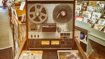 L'image montre un équipement audio vintage, un magnétophone à bande TEAC A-2300X, qui est posé sur un meuble en bois. Ce magnétophone a un design classique, avec un boîtier en métal argenté et des bobines de bande visibles sur le dessus. Deux compteurs analogiques sont situés en bas, indiquant les niveaux audio. À côté, on peut voir deux albums vinyles : l'un est intitulé "Bing & Satchmo", qui met en avant une collaboration entre le célèbre chanteur Bing Crosby et Louis Armstrong. L'autre album, "Mambo Mania" de Pérez Prado, suggère une atmosphère festive et rythmée. À l'arrière-plan, on aperçoit des étagères avec d'autres disques vinyles, créant une ambiance nostalgique et musicale.