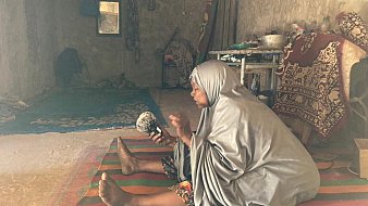 L'image représente une femme assise sur un tapis coloré dans un intérieur simple et rustique. Elle porte un vêtement ample et gris, qui couvre son corps. Sa posture est attentive, et elle tient un petit objet dans ses mains, probablement un instrument de musique ou une pièce d'artisanat. L'environnement autour d'elle est dépouillé, avec des murs en béton et quelques éléments de décoration, comme des tapis et des coussins. On peut sentir une atmosphère calme et intime, presque spirituelle. Des rayons de lumière pénètrent à travers une fenêtre, illuminant doucement la scène.