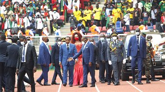 L'image montre un événement en extérieur, probablement un stade rempli de spectateurs. Au premier plan, un groupe de personnalités, dont plusieurs hommes en costume sombre et cravate, marche avec assurance. Parmi eux, une femme se distingue grâce à sa robe rouge éclatante et sa coiffure volumineuse. Ils semblent être en route pour un moment important, entourés de sécurité. À l'arrière-plan, on peut deviner une foule colorée et animée, avec des supporters portant divers vêtements aux couleurs vives, témoignant d'une atmosphère festive. L'ambiance générale semble être celle d'une grande célébration ou d'une cérémonie officielle.