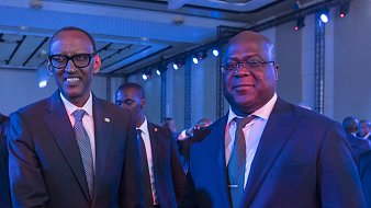 L'image montre deux hommes en costume qui se tiennent debout, se serrant la main dans un contexte de réunion ou d'événement officiel. L'un porte des lunettes et semble d'origine rwandaise, tandis que l'autre est un homme robuste, légèrement plus âgé, probablement d'origine congolaise. Ils affichent des expressions souriantes et amicales, dans une ambiance éclairée par des lumières bleues créant une atmosphère conviviale. En arrière-plan, on peut entrevoir d'autres personnes présentes à l'événement.