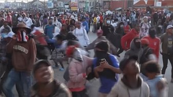 L'image montre une foule dense de personnes rassemblées dans une rue, probablement lors d'une manifestation ou d'un événement populaire. Les participants portent des vêtements variés et beaucoup d'entre eux ont des masques ou des foulards sur le visage. On peut sentir une ambiance animée et énergique, avec des mouvements rapides et des gestes expressifs. Certains individus semblent prendre des photos avec leurs téléphones. L'arrière-plan est rempli d'autres personnes, créant une atmosphère vibrante et collective.