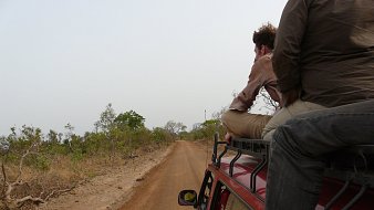 Imaginez un paysage d'arrière-pays, où un chemin de terre étroit s'étend devant vous. Sur les côtés, on peut voir de petites plantes et des buissons épars, témoignant d'une nature sauvagement préservée. La route est poussiéreuse et légèrement sinueuse, suggestive d'une aventure en milieu rural. Deux personnes sont assises sur le toit d'un véhicule, l'une d'elles se penche en arrière, le regard perdu au loin. La scène évoque un sentiment de liberté et d'exploration. L'air est probablement chargé de chaleur et du bruit léger de la nature environnante. Les couleurs sont chaudes, avec des teintes de rouge et de brun du sol, et un ciel lumineux qui crée une ambiance de voyage au cœur de la nature.
