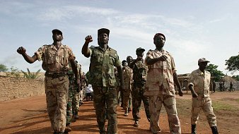 L'image montre un groupe de soldats marchant en formation sur un sol poussiéreux. Ils portent des uniformes militaires variés, allant du camouflé au beige, qui suggèrent une ambiance militaire. Certains soldats brandissent le poing en signe de détermination, tandis que d'autres avancent d'un pas ferme. En arrière-plan, on peut voir des tentes militaires ou des structures rudimentaires, suggérant un camp. Le ciel est légèrement nuageux, laissant passer quelques rayons de soleil. L'atmosphère générale évoque la discipline et la camaraderie au sein de ce groupe.
